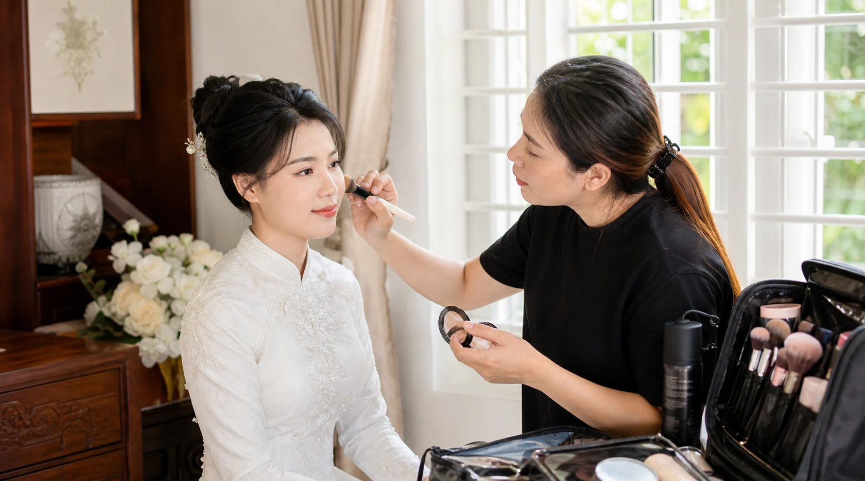 Cô dâu được trang điểm tại nhà bên cửa sổ với người makeup và bộ đồ nghề